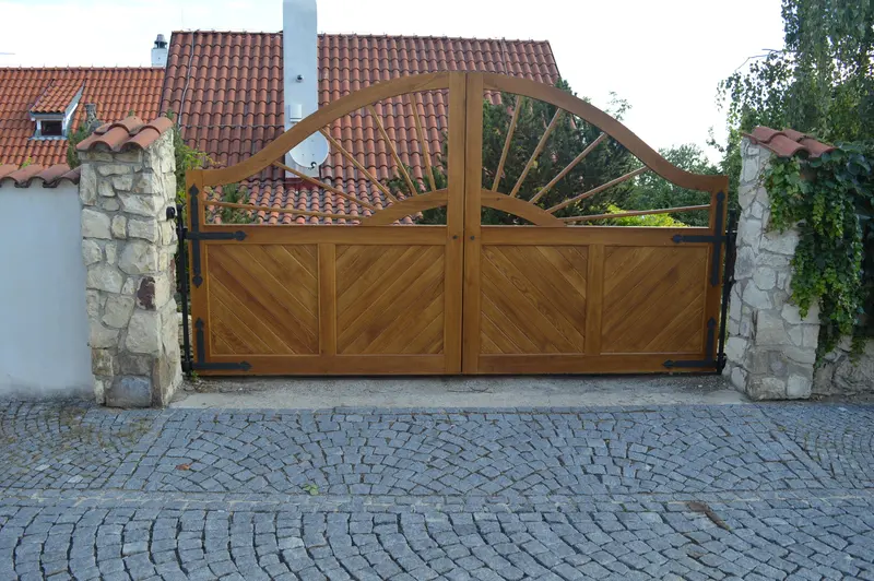 Dvoukřídlá dřevěná vjezdová brána s diagonálním vzorem a obloukovým paprskovým nástavcem mezi kamennými sloupky